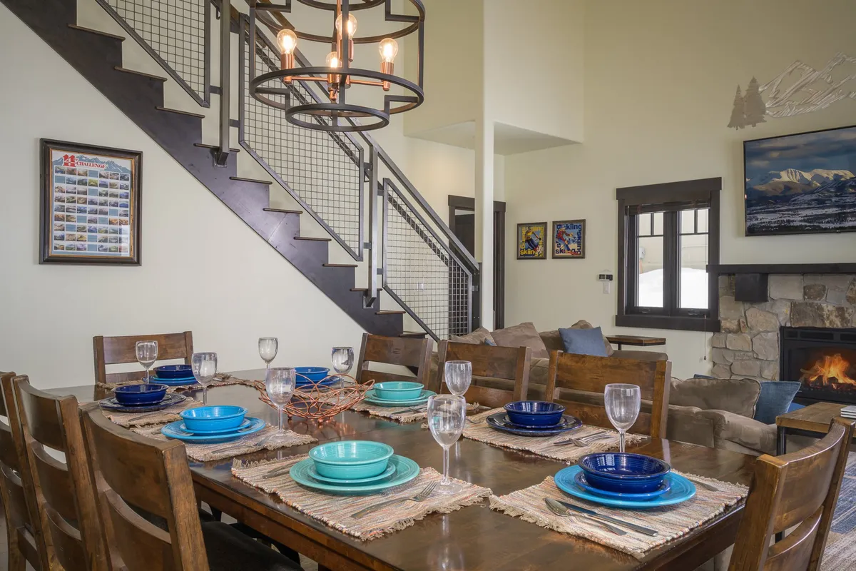 A spacious dining area with a custom live-edge wood table as the centerpiece, illuminated by a chandelier and flanked by a staircase and a cozy fireplace. The setting is ideal for shared meals, celebrations, and board games.
