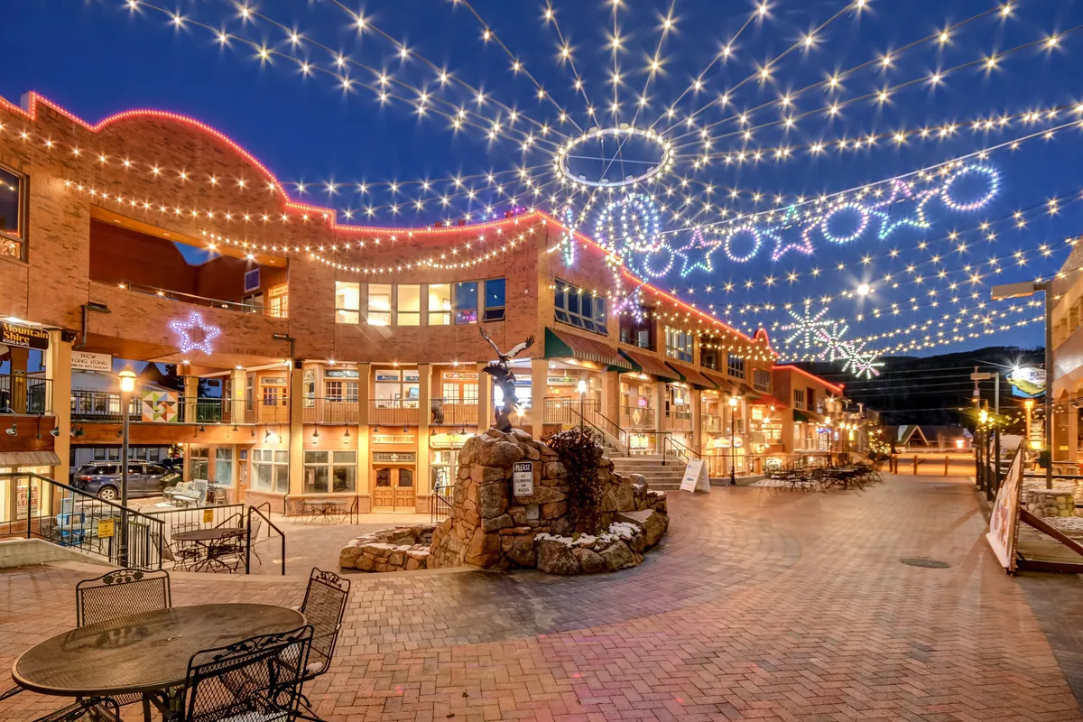 This exterior image captures a lively village scene with string lights overhead, a central sculpture, brick pavement, and inviting storefronts. It conveys a sense of joyful community and the availability of fine dining, shopping, and events within easy reach.