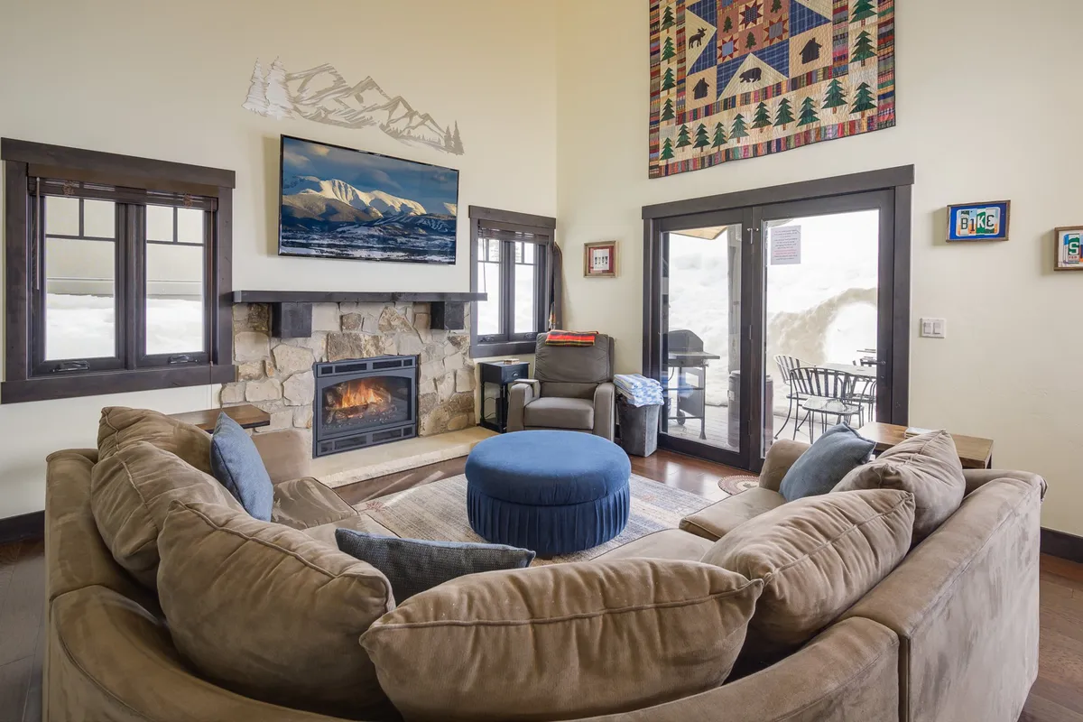 This image captures a spacious great room with a large sectional sofa facing a striking stone fireplace. Floor-to-ceiling windows frame stunning mountain views, while tasteful wall art and an adjacent outdoor seating area add to the sense of space and comfort. The atmosphere is warm and inviting, ideal for gatherings or relaxing evenings.
