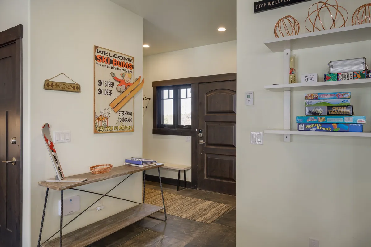 The entryway interior displays ski-themed wall art, a wooden bench for storing gear or greeting guests, and a shelf stocked with board games. The front door anchors the scene, giving a sense of arrival and anticipation for both winter sports and indoor family fun.