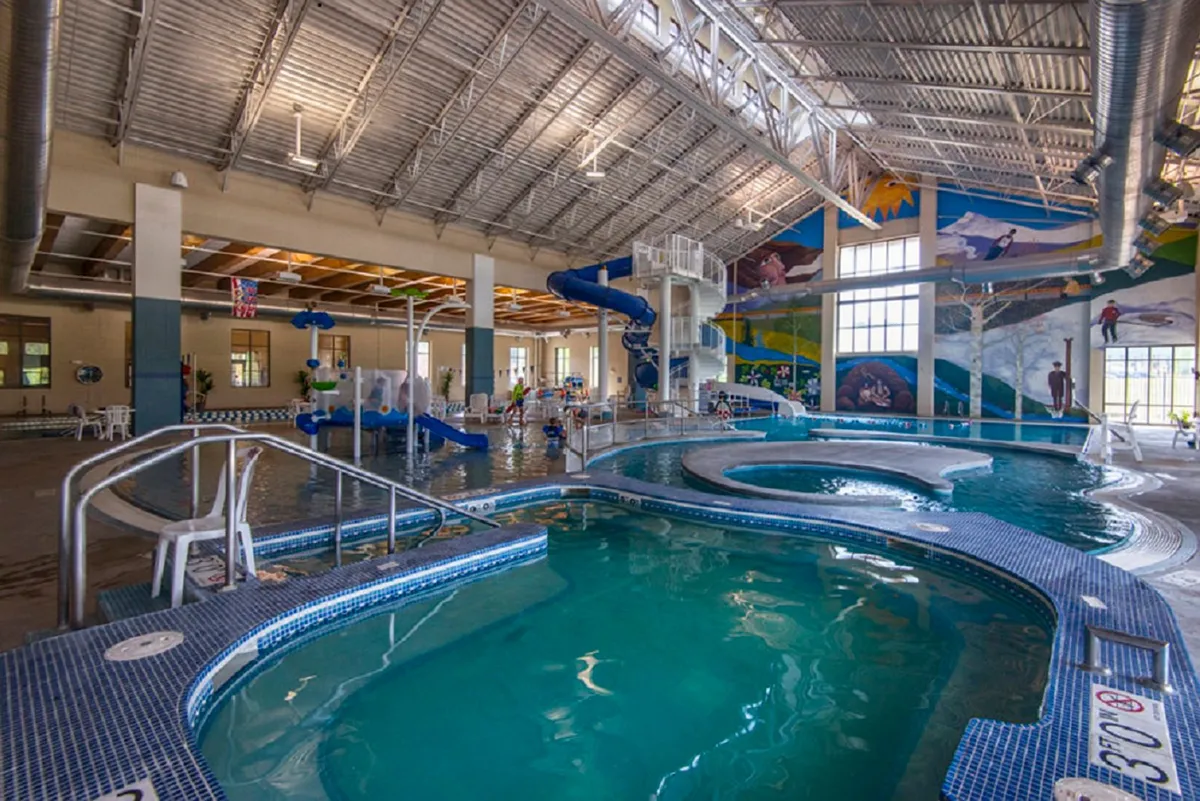 A vibrant pool area featuring colorful water slides, a spacious swimming pool, a playful mural, and children enjoying themselves. The lively atmosphere signals all-ages fun and the abundance of nearby recreational options, echoing the amenities offered at the Grand Park Rec Center.