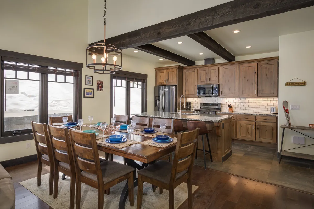 This image features a spacious, sunlit open-concept kitchen and dining area, with a large table ready for gatherings, a modern kitchen island perfect for meal prep, and expansive windows framing scenic mountain views. The warm wood tones and inviting ambiance set the stage for memorable family meals and celebrations.