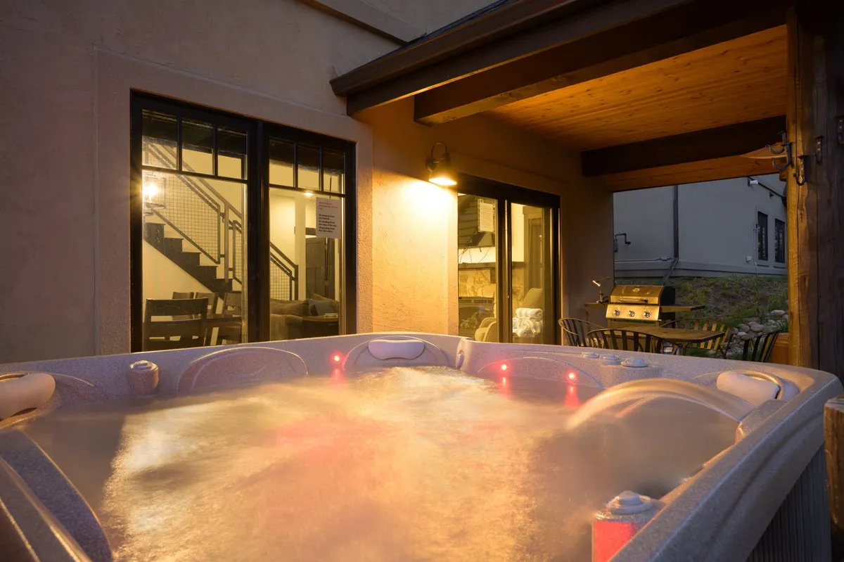 A cozy outdoor space featuring a private hot tub and a comfortable seating area, illuminated by warm interior lighting visible through large windows. The setting is ideal for relaxing after a day full of adventure, with ample space for socializing, dining, or simply soaking under the stars.